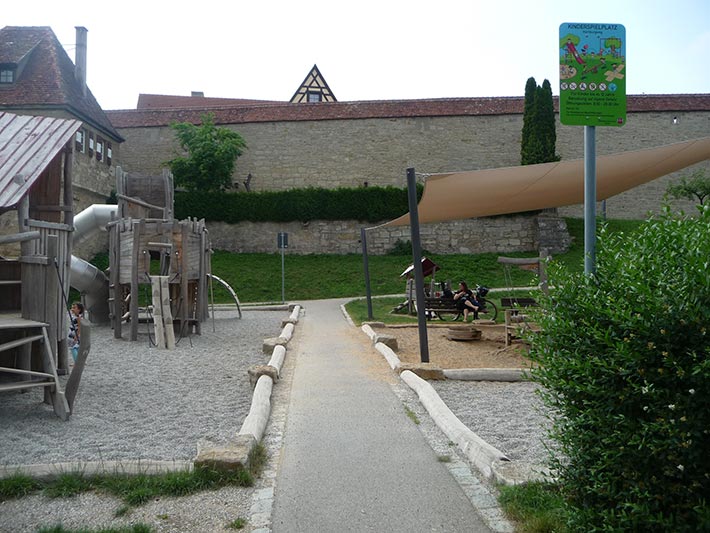 Spielplatz in Rothenburg mit Sonnensegel und Robinienstaemmen, Sitzsteinen und Fußweg.